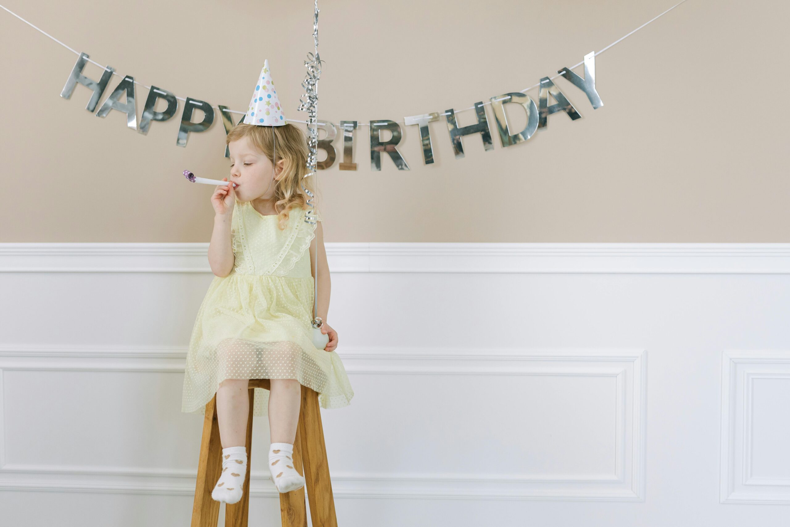 A Girl in Yellow Dress Sitting on a Wooden Stool. birthday decor in the back